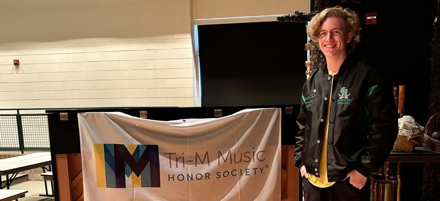 Senior Benjamin Foster, member of Tri-M's leadership team, standing next to a Tri-M banner.