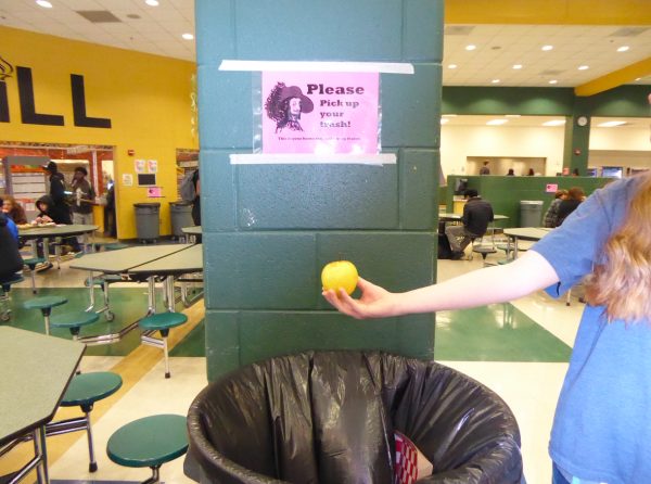 A student posing for a photo by holding an apple over a trash can in the cafeteria