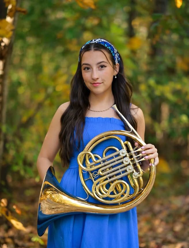 Sadie Peterson standing in front of a wooded area holding her french horn